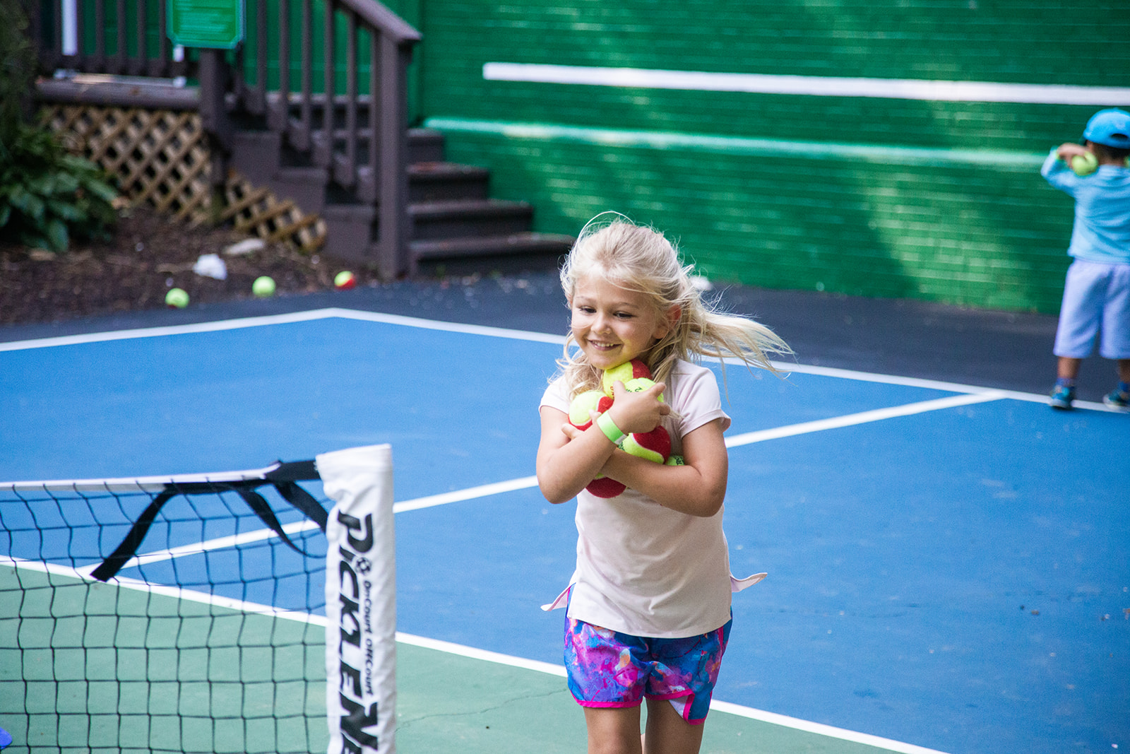 Photo of a smiling young blonde girl on an outdoor tennis court at Hollin Meadows Swim and Tennis Club holding armfuls of tennis balls.