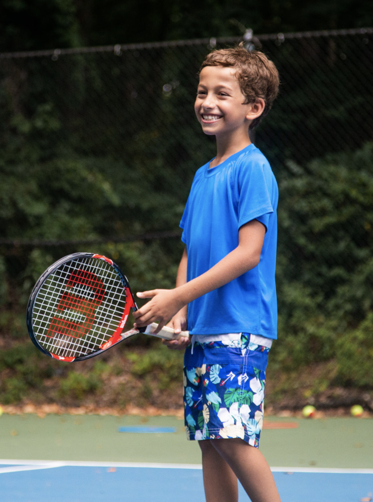photo of a young boy wearing blue, holding a tennis racket at tennis summer camp.
